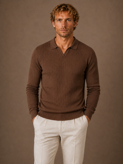 A man wearing a brown ribbed long-sleeve polo-neck sweater and off-white trousers, standing against a solid brown studio background.